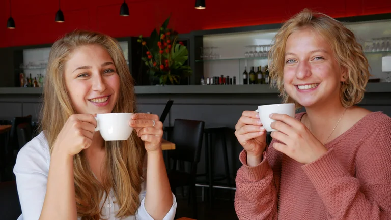 Zwei junge Frauen trinken Kaffee und schauen in die Kamera.