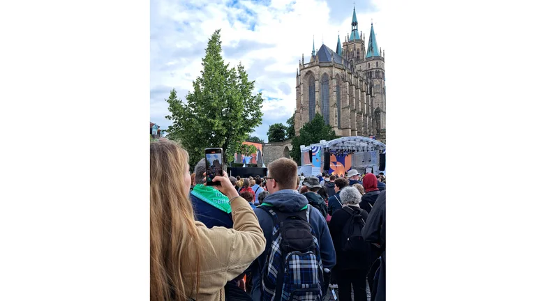 Man sieht eine Frau von hinten in einer Menschenmenge. Sie fotografiert mit ihrem Smartphone den Erfurter Dom.