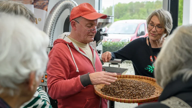 Mann zeigt Kaffeebohnen auf einer Art geflochtenem Teller. Um ihn herum schauen und h&ouml;ren Leute aufmerksam zu.