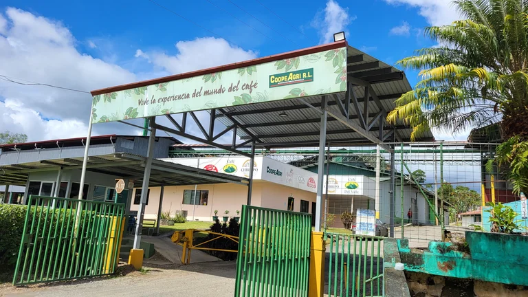 Eingang einer Kaffeekooperative mit einem gro&szlig;en Schild, auf dem &bdquo;Vive la experiencia del mundo del caf&eacute;&ldquo; und &bdquo;CoopeAgri R.L.&ldquo; steht. Das gr&uuml;ne Tor ist ge&ouml;ffnet, dahinter sind Geb&auml;ude mit Logos verschiedener Kaffeemarken zu sehen. Der Himmel ist blau mit wei&szlig;en Wolken.
