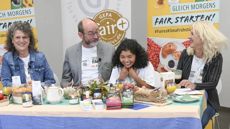 Drei Frauen und ein Mann sitzen an einem &uuml;ppig gedeckten Fr&uuml;hst&uuml;ckstisch mit vielen GEPA-Produkten und lachen ausgelassen. Hinter ihnen sind GEPA-Banner aufgestellt mit der Aufschrift "Gleich morgens fair starten!"