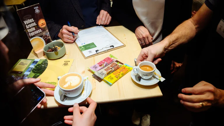 Ein Tisch mit den drei neuen Nussriegeln, mehreren Kaffeetassen und Arbeitsunterlagen, darum herum sitzen Personen, deren H&auml;nde zu sehen sind.