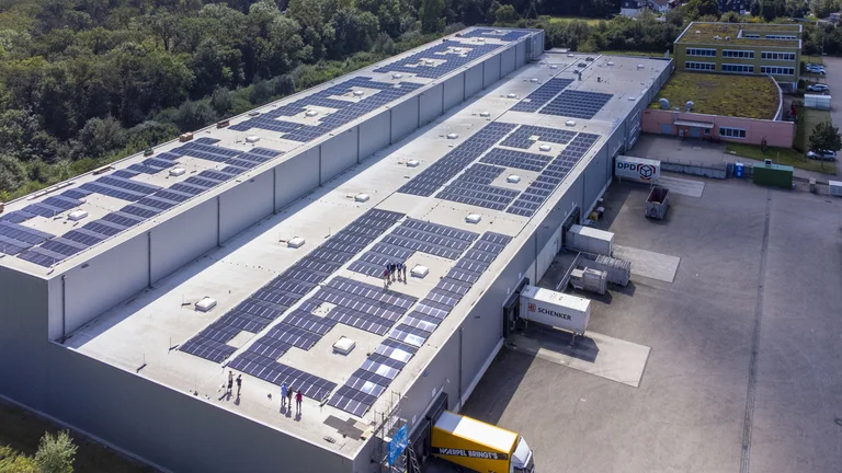 Drohnenaufnahme vom Lagerdach der GEPA von schräg rechts: Über das ganze Dach verteilt sind in Reihen angeordnet Solarpaneele zu sehen, außerdem laufen ein paar Menschen darauf herum, die winzig wirken.