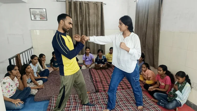 Mann und Frau stehen sich in einem Selbstverteidigungskurs gegen&uuml;ber. Sie simuliert einen Angriff auf ihn. Um sie herum sitzen ca. 15 M&auml;dchen auf Kissen und sehen zu.