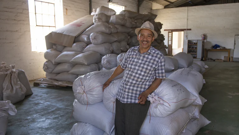 Mann mit Hut in einer Lagerhalle lehnt an einem Stapel weißer Säcke. Im Hintergrund sind weitere Säcke gestapelt.