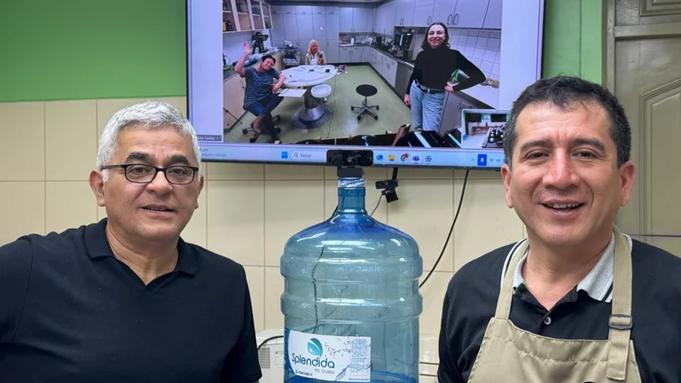 Zwei M&auml;nner stehen links und rechts von einem Wasserspender. Hinter ihnen an der Wand ist ein Bildschirm. Darauf sind per Videocall drei Menschen aus einem Kaffeelabor zugeschaltet.