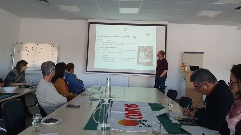Sechs Personen an einem Tisch in einem Tagungsraum lauschen einer Veranstaltung. Vorne steht ein Mann vor einer Leinwand und präsentiert. Gerade ist eine Folie mit der Überschrift "O que significa Fair Plus na GEPA?" zu sehen.