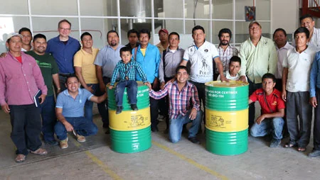 22 Personen in einer Art Fabrikgeb&auml;ude schauen in die Kamera. Sie stehen um zwei gr&uuml;n-gelb-gr&uuml;ne F&auml;sser.