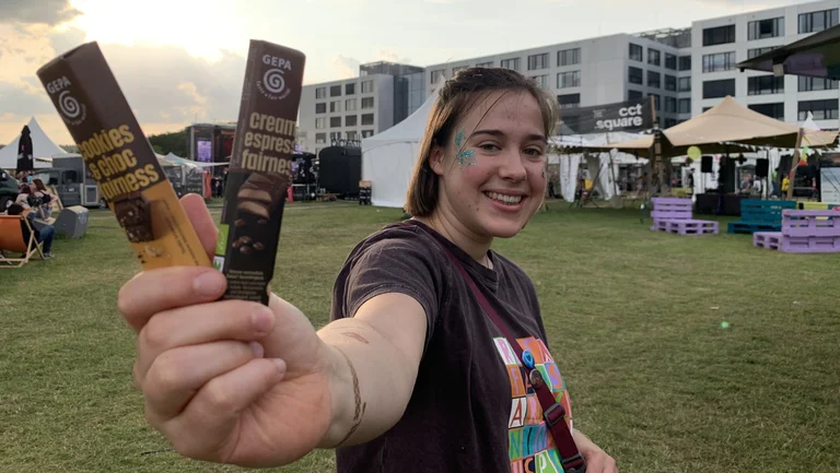 Junge Frau mit Glitzer im Gesicht steht auf einem Festivalgel&auml;nde. Sie h&auml;lt zwei GEPA-Schokoriegel in die Kamera und l&auml;chelt.