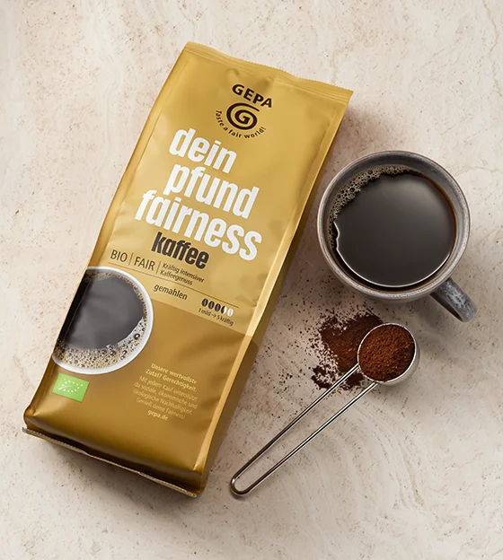 Eine Packung GEPA-Kaffee &bdquo;dein pfund fairness&rdquo; arrangiert mit einer Tasse Kaffee und einem L&ouml;ffel Kaffeepulver.