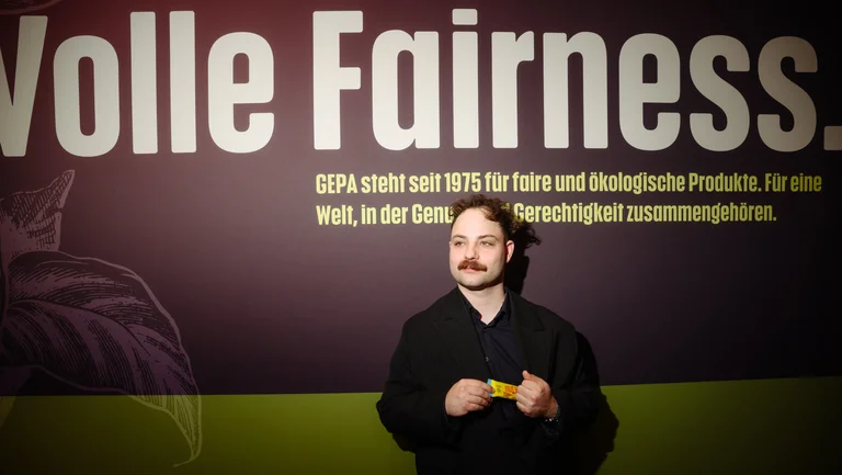 Junger Mann steht vor Plakat mit Slogan &ldquo;Volle Fairness&rdquo; und h&auml;lt einen GEPA-Nussriegel &ldquo;Mango Cocos&rdquo;