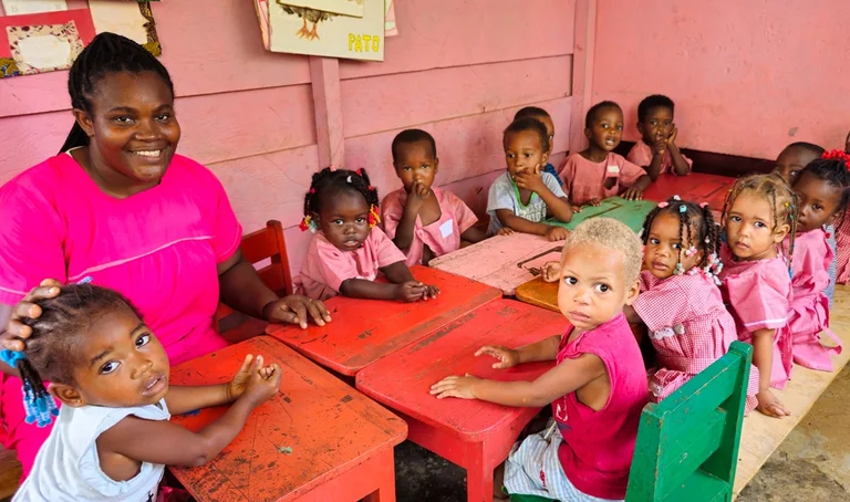Eine Kinderg&auml;rtnerin sitzt mit Kindergartenkindern an Tischen. Sie blicken in die Kamera.