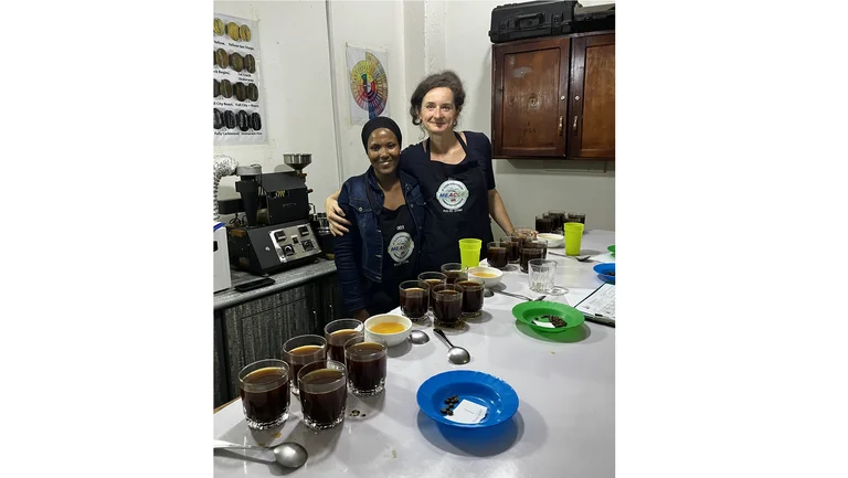Zwei Frauen an einem Tisch mit aufgegossenen Kaffeeproben, Kaffeezubeh&ouml;r, im Hintergrund eine Kaffeemaschine sowie ein Aromen-Poster.