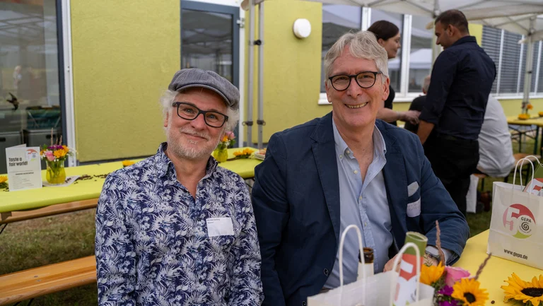 Zwei Herren mit Brille sitzen an einer dekorierten Biergarnitur und lächeln in die Kamera. Bei einem ist das Namensschild mit der Aufschrift „Michael Kaufung, Misereor“ zu erkennen.