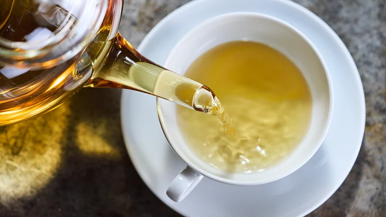 Aus einer Glaskanne wird hellgr&uuml;ner Tee in eine wei&szlig;e Tasse gegossen.