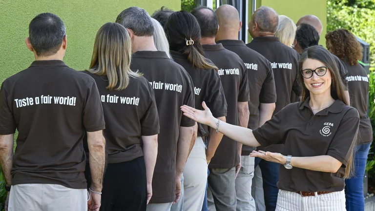 Dreizehn Erwachsene stehen mit dem R&uuml;cken zur Kamera. Auf ihren braunen T-Shirts steht "Taste a fair world!" Eine junge Frau im braunen T-Shirt mit GEPA-Logo l&auml;chelt in die Kamera und deutet auf den Schriftzug auf den T-Shirts der anderen.
