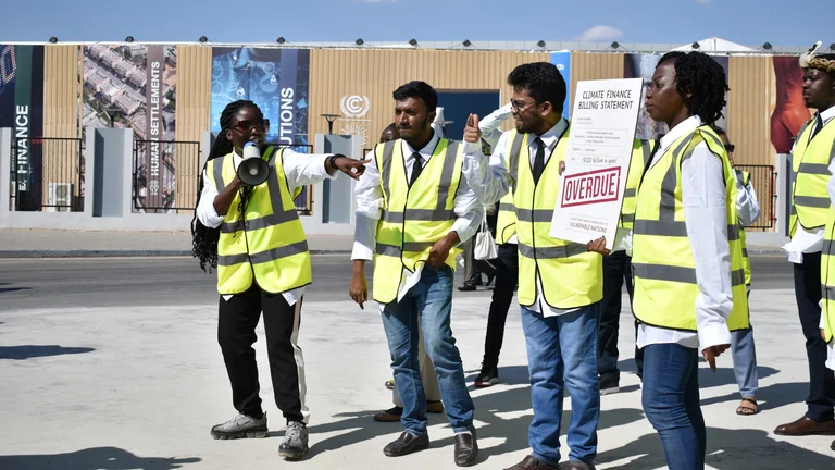 Zwei Frauen und zwei Männer in gelben Warnwesten stehen vor einem Gebäude der Klimakonferenz COP 27. Eine hält ein Megafon und deutet auf ein Plakat, das ein anderer hält. Darauf steht: "Climate Finance Billing Statement. OVERDUE!"
