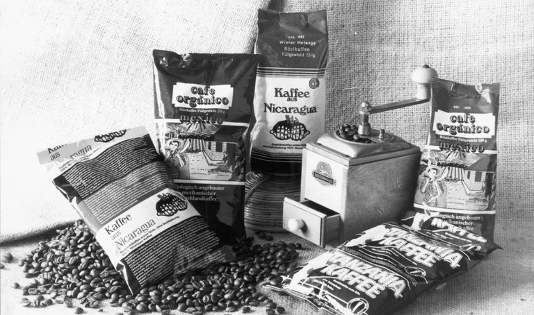 Schwarz-Weiß-Bild: Arrangement von Packungen mit fair gehandeltem Kaffee auf Kaffeebohnen. Dazwischen steht eine alte Kaffeemühle