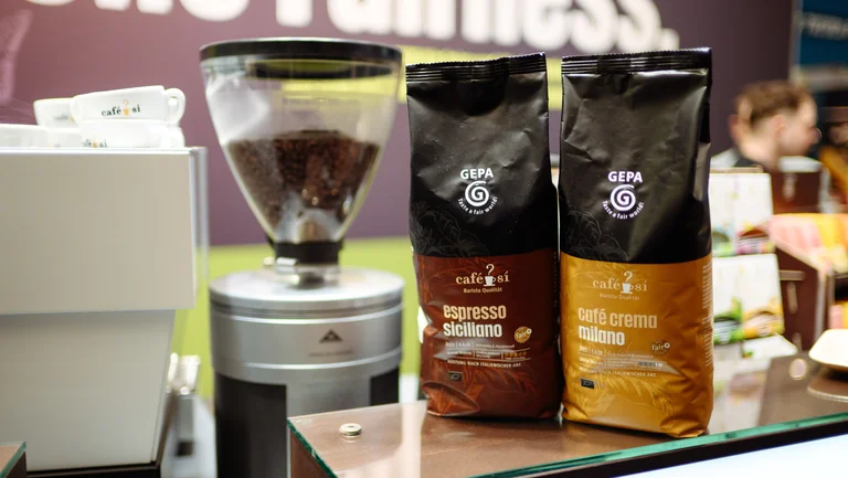 Zwei GEPA-Kaffeepackungen der Barista-Varianten &ldquo;espresso siciliano&rdquo; und &ldquo;caf&eacute; crema milano&rdquo; stehen neben einer gef&uuml;llten Kaffeem&uuml;hle.