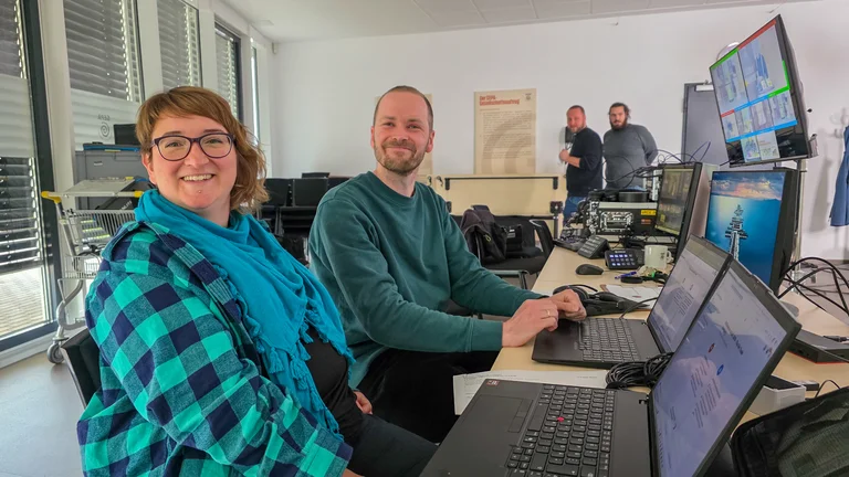 Lukas N&ouml;th und Anne Bien aus der GEPA-Onlineredaktion sitzen an ihren Laptops und lachen in die Kamera. Im Hintergrund sind weitere Technik-Aufbauten sowie das Team vom Rheinklang Media zu sehen. 