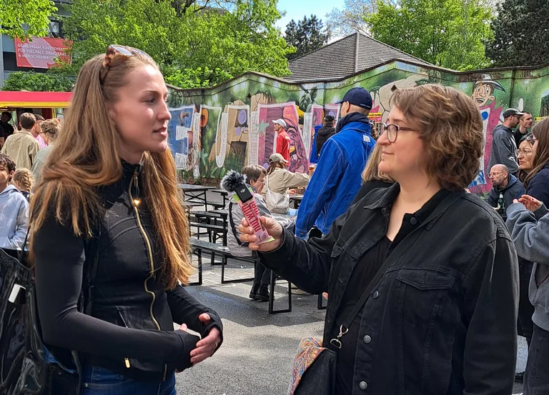 Zwei Frauen im Gespr&auml;ch, eine h&auml;lt ein Mikrofon. Im Hintergrund Festivaltreiben und Grafitti. 