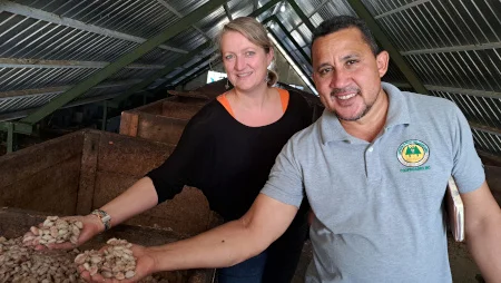 Frau und Mann stehen unter einem niedrigen Dach und halten frisch entnommene Kakaobohnen in Händen. Sie lächeln in die Kamera.