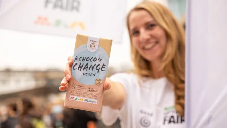 Junge Frau auf einer Veranstaltung hält eine GEPA-Klimaschokolade #Choco4Change Vegan in die Kamera und lächelt.