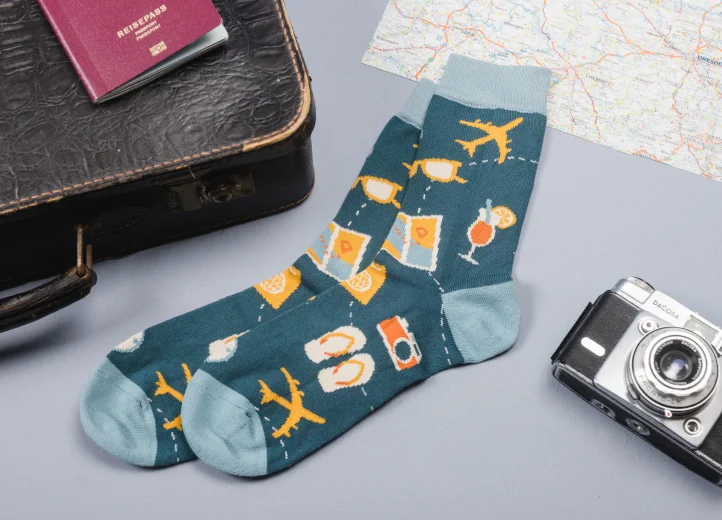 Ein Paar Socken mit bunten Reisemotiven darauf. Neben ihnen liegen ein Koffer, ein Reisepass, eine Kamera und eine Landkarte.