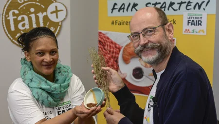 Frau und Mann l&auml;cheln in die Kamera. Sie h&auml;lt eine Schale mit etwas getrocknetem Rooibos-Tee darin, er einen Rooibos-Zweig. Hinter den beiden sind GEPA-Banner aufgestellt.