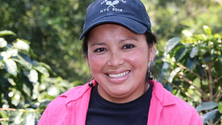 Portrait einer Frau mit Kappe. Sie steht vor Kaffeepflanzen und l&auml;chelt in die Kamera.