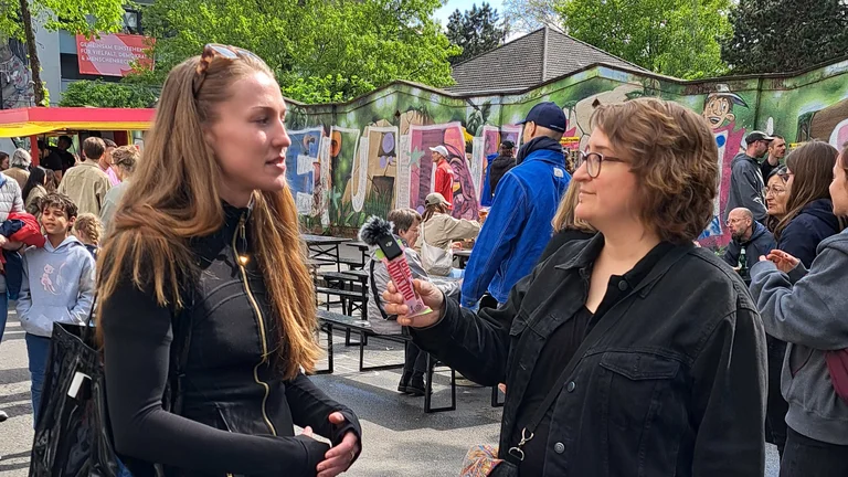 Zwei Frauen im Gespr&auml;ch, eine h&auml;lt ein Mikrofon. Im Hintergrund Festivaltreiben und Grafitti. 