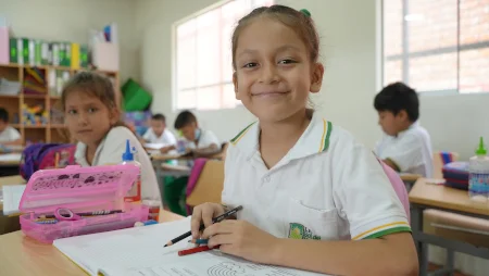M&auml;dchen sitzt an ihrem Platz in einem Klassenzimmer Schultisch und l&auml;chelt in die Kamera. Vor ihr ein aufgeschlagenes Heft und ein M&auml;ppchen