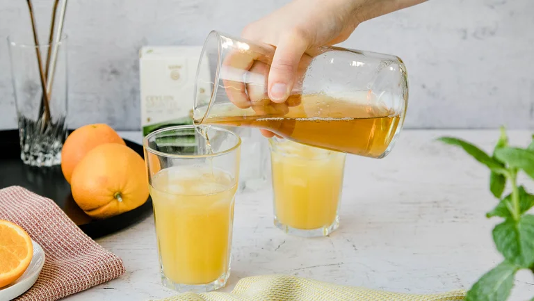 Zwei Gläser mit gelber Limonade darin, daneben sind ein Tuch, eine Schale mit Orangen und eine Limonadenflasche platziert