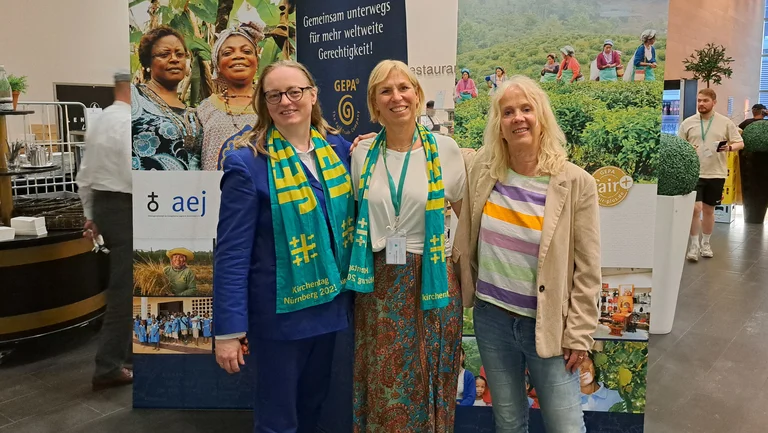 Drei Frauen haben die Arme &uuml;ber ihre Schultern gelegt und schauen in die Kamera. Sie stehen vor Bannern in einer Messehalle.