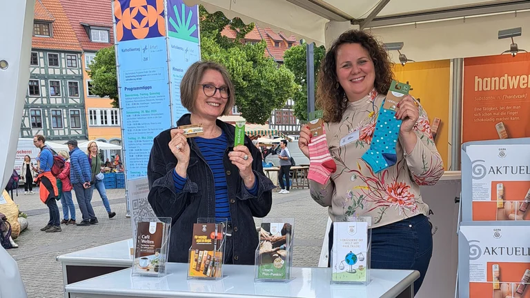 Zwei Frauen an einem Messestand mit ausgelegtem Infomaterial. Sie halten Socken bzw. einen Sesamriegel in die Kamera.