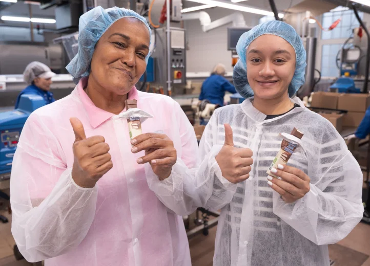 Zwei Frauen mit Haarnetz und Kitteln in einer Schokoladenfabrik präsentieren Schokoriegel und halten Daumen hoch