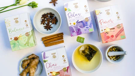 Blick von oben auf arrangierte Packungen GEPA-Tee. Dazwischen liegen eine Ingwerknolle , Sternanis in einer Schale, Zimtstangen, ein mit Tee und Beutel gef&uuml;llte Tasse und ein M&ouml;rser.