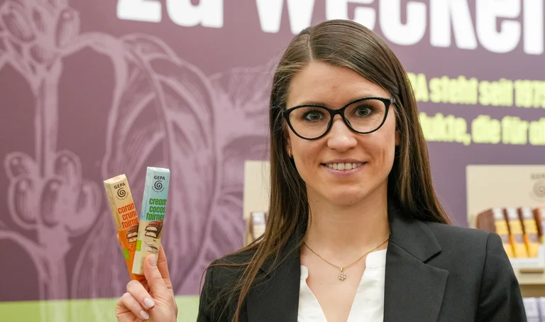 Junge Frau mit Blazer und Brille hält an einem Messestand zwei GEPA-Riegel in der Hand