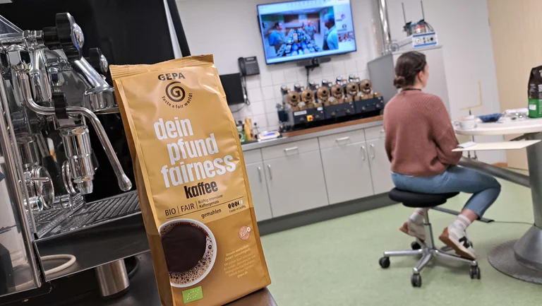 In einem Kaffeelabor: Auf einem Tresen im Vordergrund steht eine Packung GEPA-Kaffee "dein pfund fairness". Im Hintergrund sitzt eine junge Frau an einem Tisch. Auf einem Bildschirm an der Wand sieht man drei M&auml;nner, die sich ebenfalls in einem Kaffeelabor befinden.