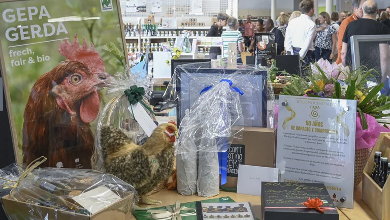 Tisch mit Geschenken, einer Urkunde und einem Plakat mit einem Huhn darauf. Darauf steht „GEPA GERDA. Frech, fair & bio“ Im Hintergrund sind Menschen auf einer Veranstaltung zu sehen.