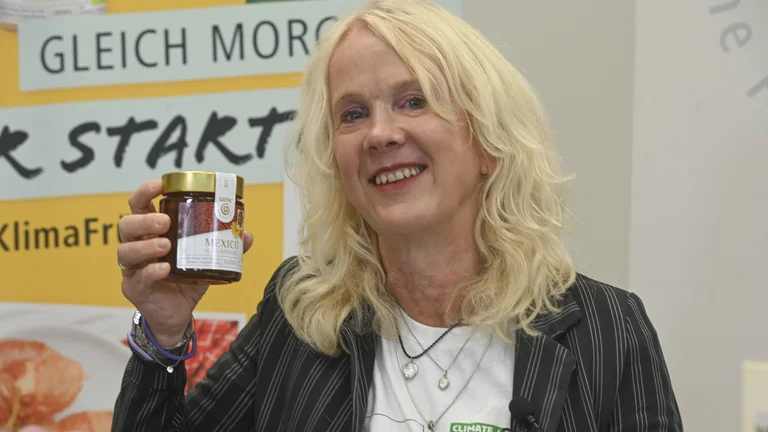 GEPA-Pressesprecherin Barbara Schimmelpfennig l&auml;chelt in die Kamera und h&auml;lt ein Glas GEPA-Honig hoch. Hinter ihr ein GEPA-Banner mit der Aufschrift "Gleich morgens fair starten"