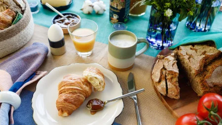 Blick auf einen bunt gedeckten Fr&uuml;hst&uuml;ckstisch mit Croissant auf einem Teller, Marmelade, Brot, Kaffeetasse, einem Glas Orangensaft, Ei in einem Eierbecher, Blumenvase etc.