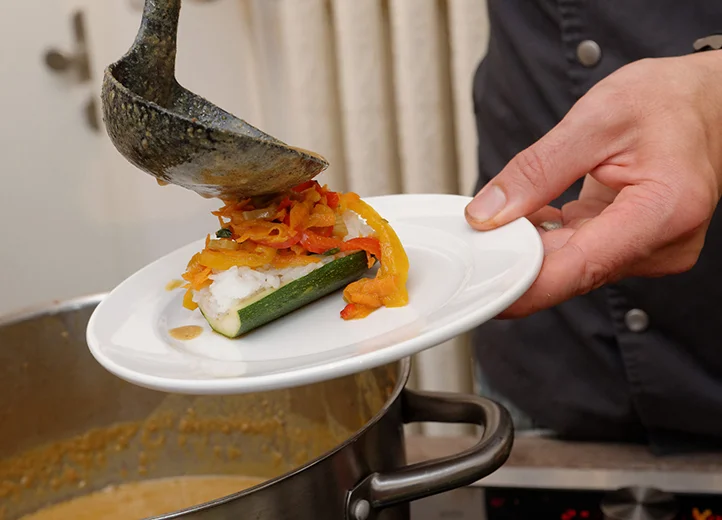 Eine Hand hält einen Teller mit einer Zucchini darauf. Aus einer Kelle kommt Gemüse darauf