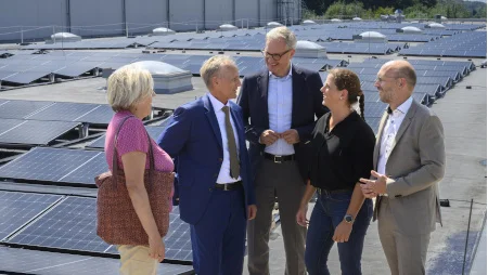Fünf Menschen stehen im Halbkreis auf einem Flachdach mit vielen Solarpaneelen und unterhalten sich.