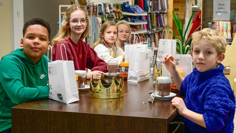 F&uuml;nf Kinder sitzen an einem Tisch und schauen in die Kamera. Vor ihnen auf dem Tisch stehen Gl&auml;ser mit Kakao, wei&szlig;e Geschenkt&uuml;ten mit GEPA-Logo und der Aufschrift "echt fair" und eine goldene Krone.