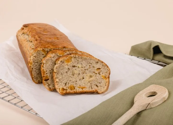 Auf weißem Papier auf einem Rost liegt ein Bananenbrot, von dem zwei Scheiben schon abgeschnitten sind. Daneben liegt dekorativ ein olivgrünes Tuch mit einem Holzlöffel darauf.