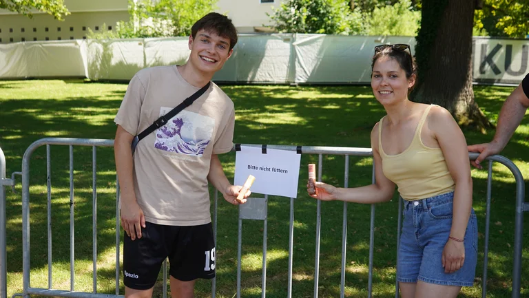 Junger Mann und junge Frau stehen vor einer mit Gitter abgesperrten Wiese. Am Gitter steht ein Schild mit "Bitte nicht f&uuml;ttern". Sie halten je einen GEPA-Schokoriegel und l&auml;cheln in die Kamera.