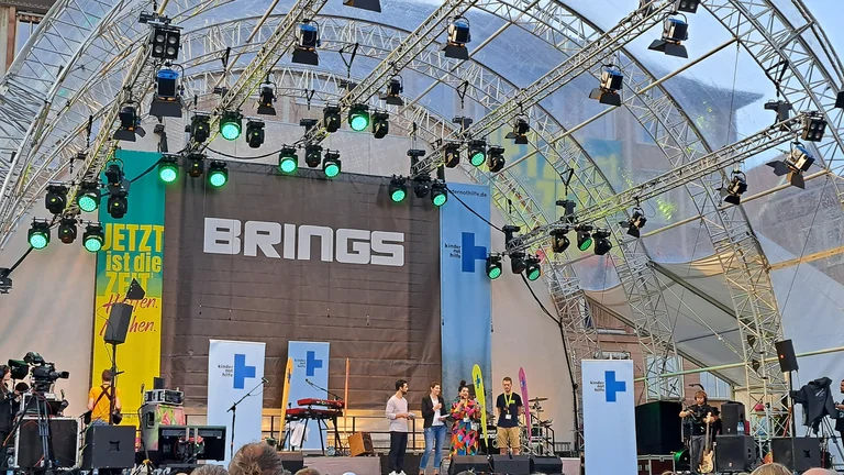 Vier junge Menschen mit Mikrofonen stehen auf einer gro&szlig;en B&uuml;hne. Hinter ihnen h&auml;ngt ein riesiges Banner mit dem Schriftzug "Brings".