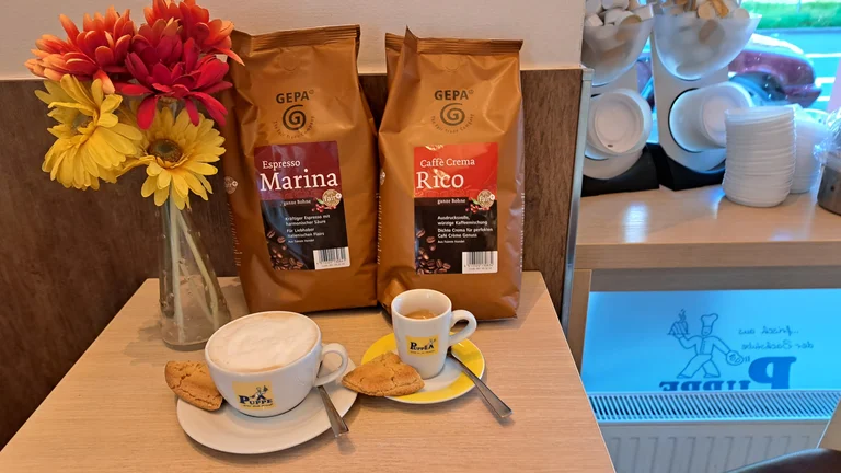 Auf einem Tisch stehen zwei Packungen GEPA-Kaffee "Marina" bzw. "Rico". Dazu Blumen in einer Vase und zwei Unterteller mit je einem Geb&auml;ckst&uuml;ck und einer Tasse Kaffee darauf.