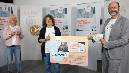 Zwei Frau und ein Mann stehen in einer Art Studio vor GEPA-Bannern. Sie halten Tafeln Schokolade sowie gemeinsam eine riesige Postkarte mit gestaltetem Motiv, u.a. mit GEPA-Logo und der Aufforderung "Mach mit!"